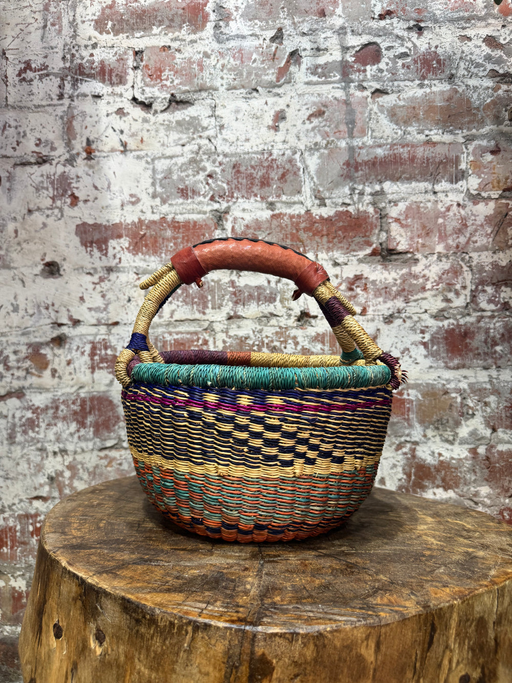 Woven basket with colorful handle on a wooden stump against a textured brick wall.