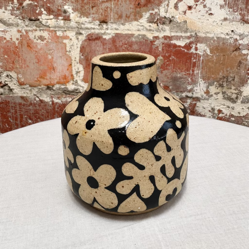 A small ceramic vase with a glossy black glaze and large beige abstract floral shapes, set on a white fabric-covered surface against a distressed brick wall.