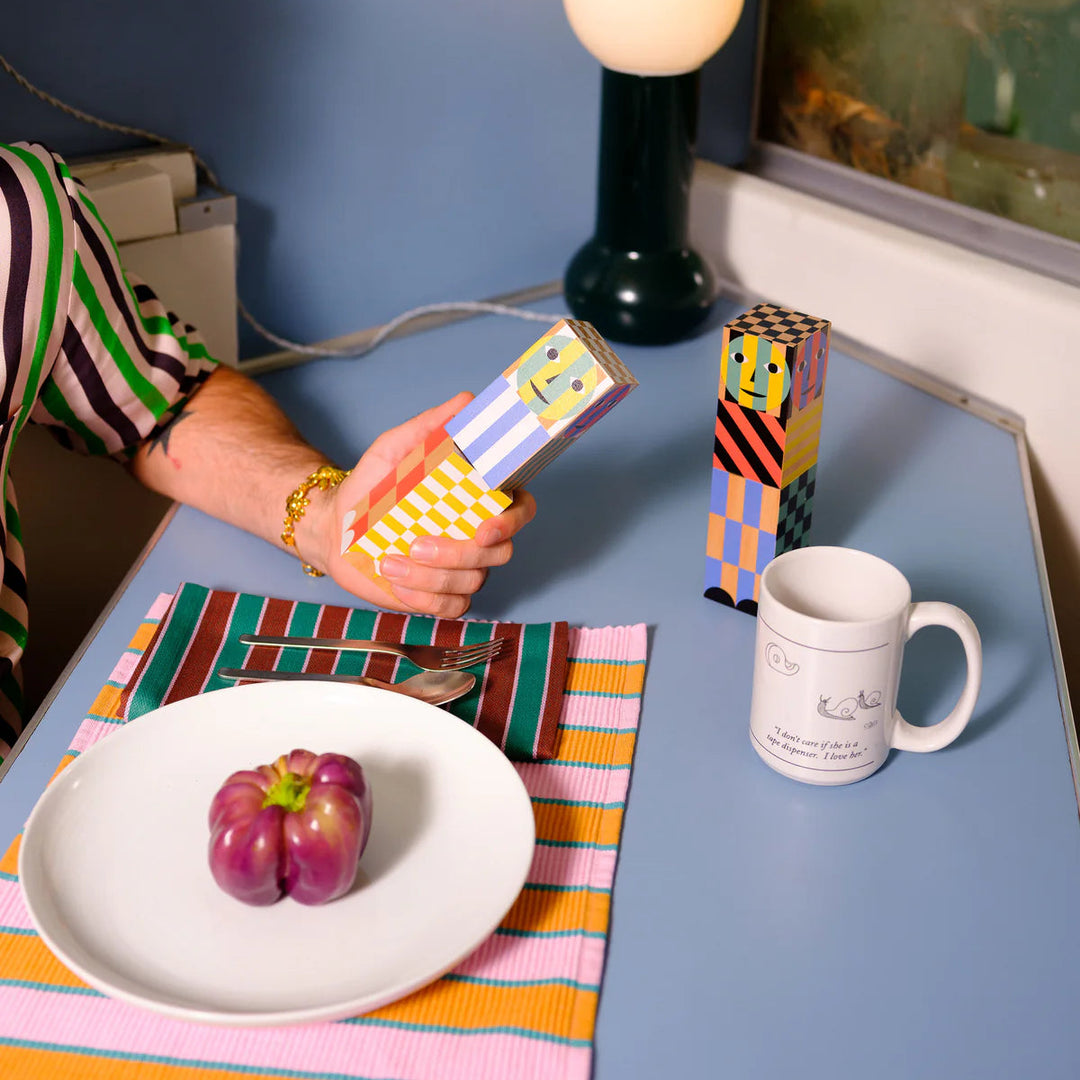 Person holding a colorful box at a table with a plate, mug, and lamp.