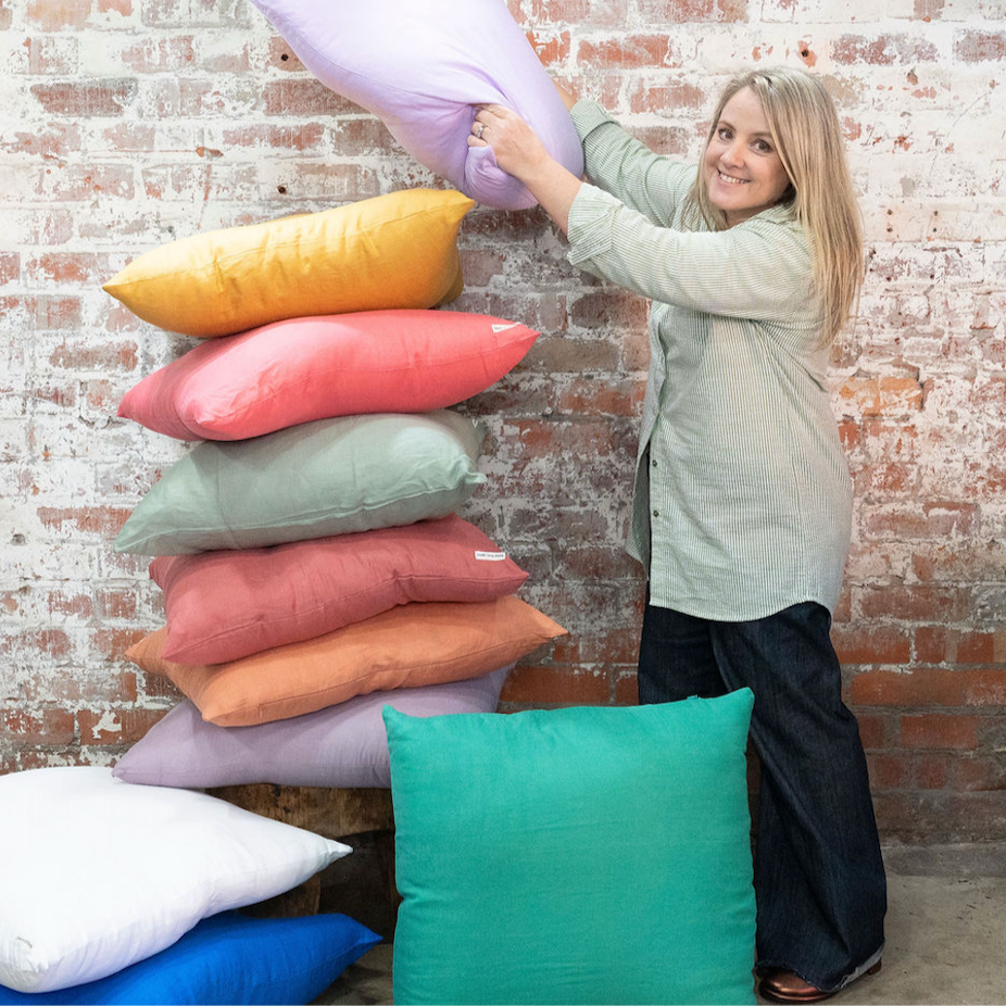 Woman arranging colorful pillows against a brick wall.