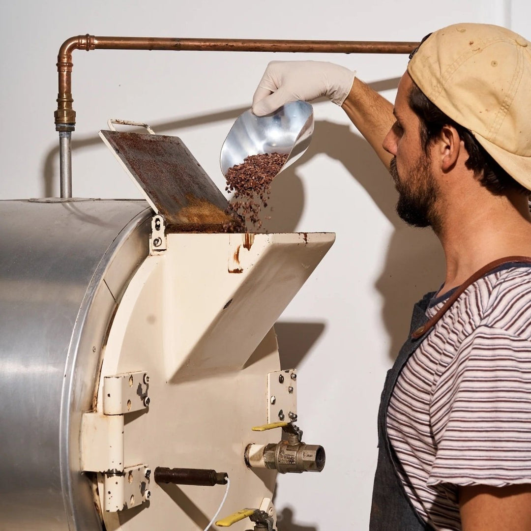 Person operating a large metal machine, likely a coffee roaster, with a white background.