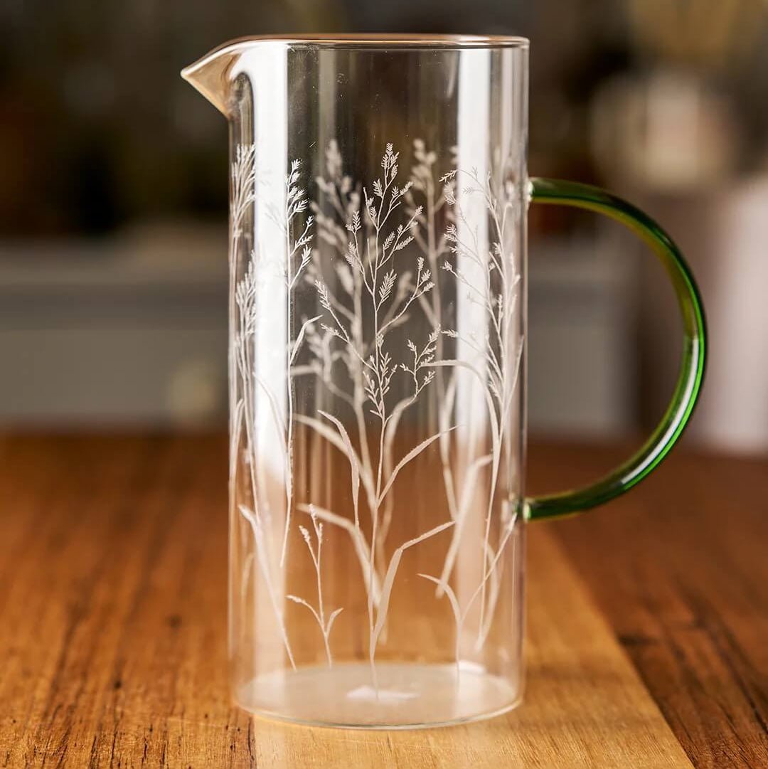 Clear glass pitcher with nature-inspired design and green handle on wooden surface
